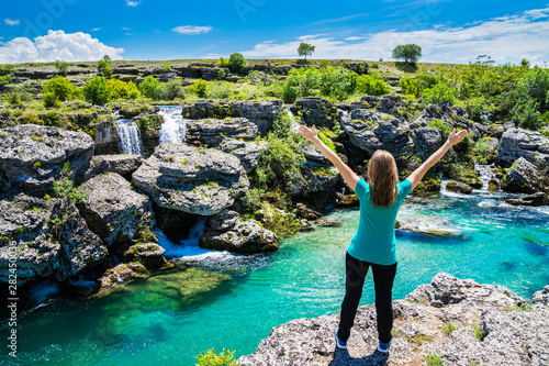 Montenegro, Pretty blonde young woman standing with arms stretched amazed at azure water of cijevna river riverside in spectacular nature landscape on sunny day