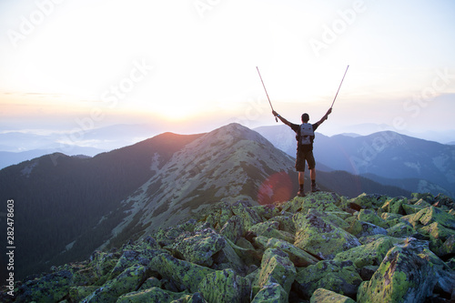 caucasian man hiker is on the peak of green stones mountain watching sunset with trekking sticks in the hands in the air
