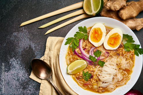 Mohinga at dark background in white plate. Mohinga is a burmese cuisine fish soup with catfish, rice noodles, chicken eggs and lime. Copy space on the left