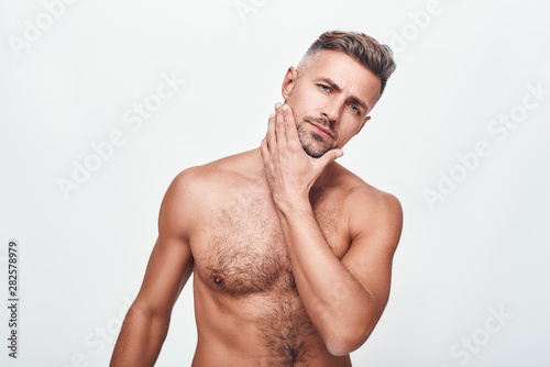 Sexy and confident. Handsome man with a stubble keeping touching his face and looking at camera while standing against grey background