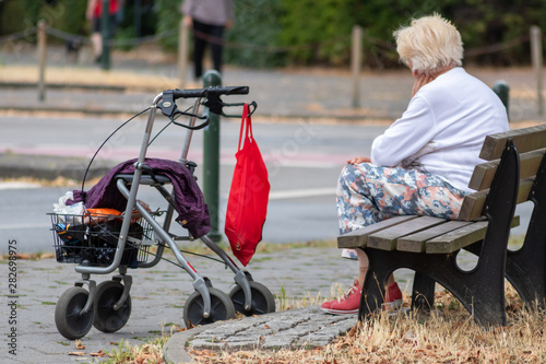Einsame alte Frau und Großmutter sitzt auf einer Bank am Straßenrand und wartet allein auf den Bus oder ein Taxi mit ihrem Rollator, um die Einkäufe in roter Tasche ins Seniorenheim zu bringen