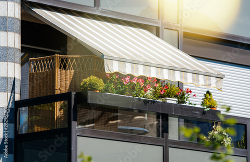 French balcony with beautiful awning and flowers covered with rays of sun - protection during hot weather and radiation