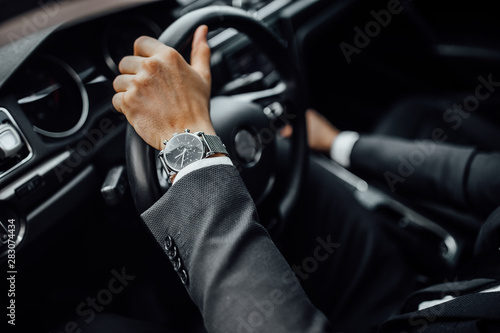 Close up top view of man's watch in black suit keeping hand on the steering wheel while driving a luxury car.