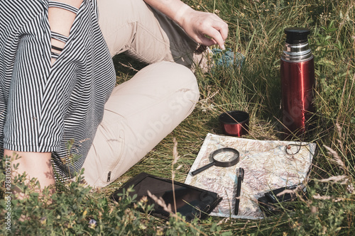 Traveler girl in a striped T-shirt and brown pants sits on the grass. Nearby lies a map, and on it is a tablet, magnifier, binoculars, pen. Nearby is a thermos with a mug.