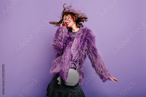 Laughing curly woman in sparkle sunglasses dancing on purple background. Happy girl in fluffy coat enjoying photoshoot in winter clothes.