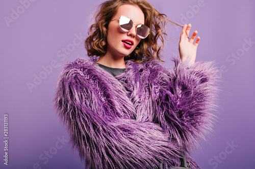 Dreamy pretty girl plays with short curly hair standing on bright background. Indoor portrait of pensive female model in sunglasses and purple fluffy coat.