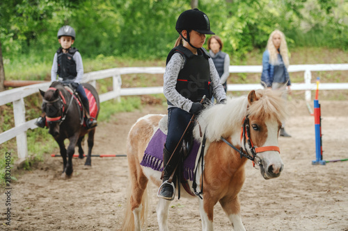 Children with helmets and protective vests on riding pony horses at sunny day on ranch.
