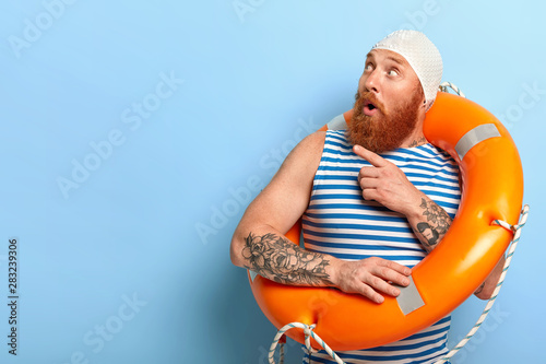 Surprised unshaven male vacationist dressed in bathingcap and striped vest, poses with swimring, points aside with index finger and looks surprisingly aside on blank space for your promotion