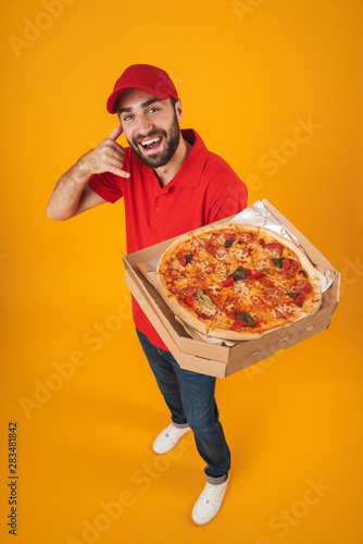 Full length image of positive delivery man in red uniform smiling and holding pizza box