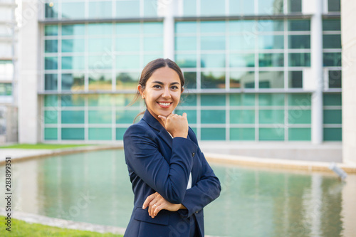 Happy confident young businesswoman posing outside. Beautiful Latin woman wearing formal suit, standing near office, leaning chin on hand and looking at camera. Successful business woman concept