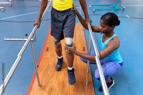 Physiotherapist assisting disabled man walk with parallel bars in sports center
