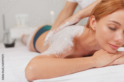 Cropped close up of a beautiful happy woman smiling with eyes closed, getting body wrap treatment at spa. Professional spa therapist applying white mud on the body of beautiful woman
