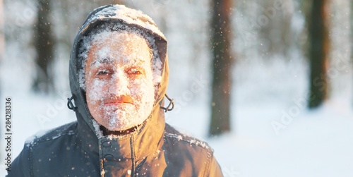 Fun portrait of an young frozen man. Jogging in a blizzard in the woods. Face covered with snow and frost. Closeup portrait of happy young guy smiles in cold weather in the winter forest at sunset.