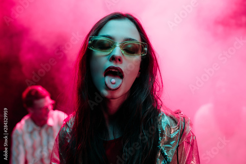 beautiful girl in sunglasses with lsd on tongue in nightclub with pink smoke