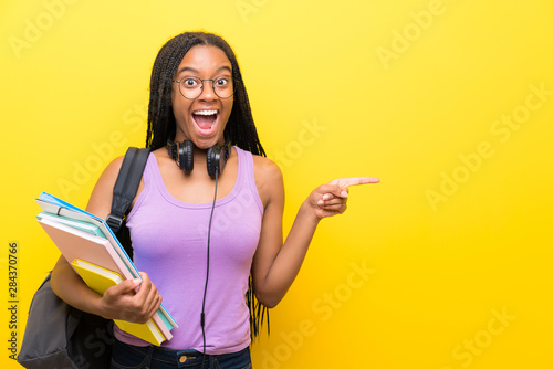 African American teenager student girl with long braided hair over isolated yellow wall surprised and pointing finger to the side