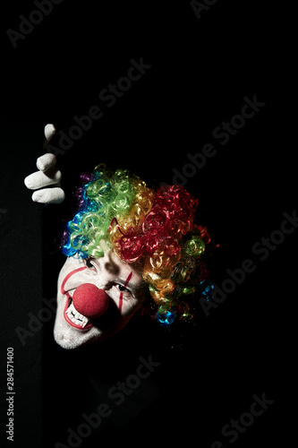 Scary clown peeping around the corner of a black wall. He is wearing a colored wig and sharp fangs.