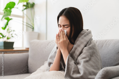 sick asian young woman sneezing into tissue paper while covered with a blanket and sitting on sofa in the living room