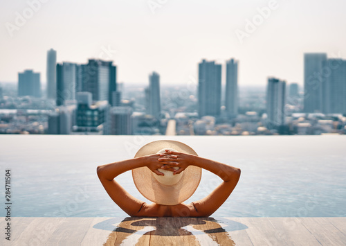 Back view of young woman in hat relaxing in swimming pool on the roof top of hotel and enjoy cityscape