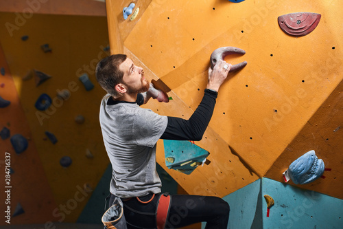 Side shot of well equipped sportsman training in colorful bouldering gym, climbing at difficult rock wall, overcomes his physical disability, has strong will and power. Sport and motivation concept.