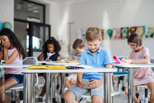 A small school boy with smartphone sitting at the desk in classroom, playing.