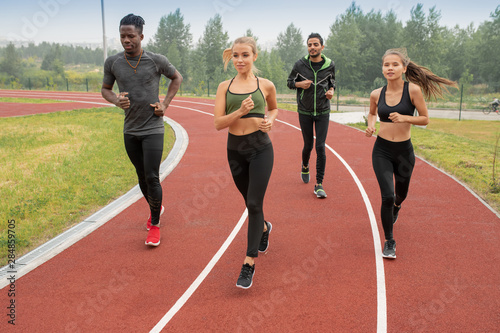Group of active guys and girls in sportswear running down racetracks on stadium