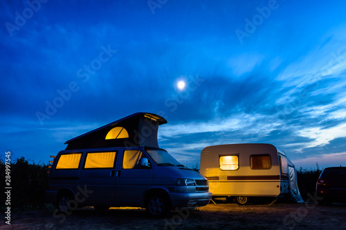 A camper van with its pop top roof open parked next to a vintage caravan in a campsite at nightfall, both illuminated from inside and the moon glowing above in a stormy sky.