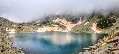 Panoramic view of Rila mountain lake on a misty, snow covered landscape with foreground rocks