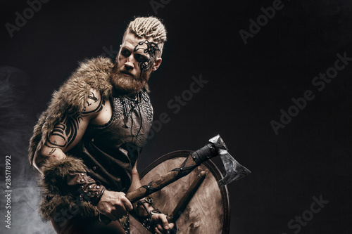 Medieval warrior berserk Viking with tattoo on skin, red beard and braids in hair with axe and shield attacks enemy. Concept historical photo