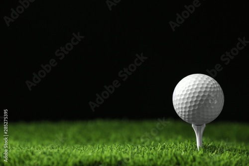 Golf ball with tee on artificial grass against black background, space for text