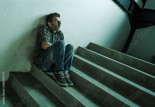 dramatic portrait of young depressed and paranoid man sitting outdoors on dark grunge street corner staircase feeling sick suffering depression problem and anxiety crisis