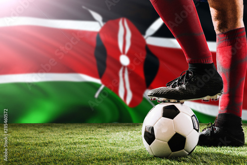 Close up legs of Kenya football team player in red socks, shoes on soccer ball at the free kick or penalty spot playing on grass.