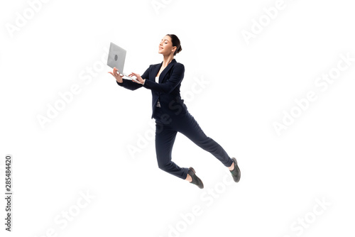 young cheerful businesswoman using laptop while levitating isolated on white