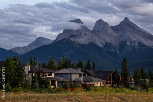 Beautiful view of Residential Homes with Canadian Rocky Mountains in the background during a cloudy summer sunset. Taken in Canmore, Alberta, Canada.