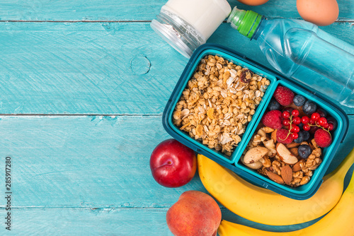 school lunch boxes with granola, vegetables, nuts, berries, banana, eggs, peaches and water on blue background