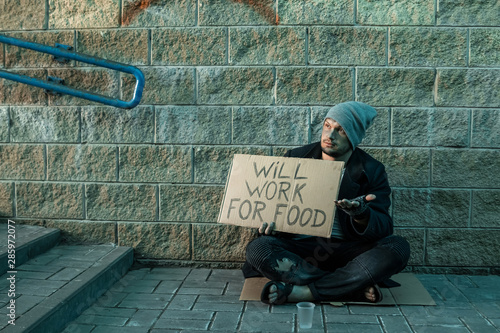 A man, homeless, a man asks for alms on the street with a sign will work for food. Concept of homeless person, addict, poverty, despair.