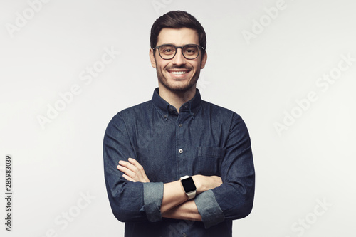 Man with crossed arms wearing glasses and smart watch, looking at camera with smile, standing isolated on gray