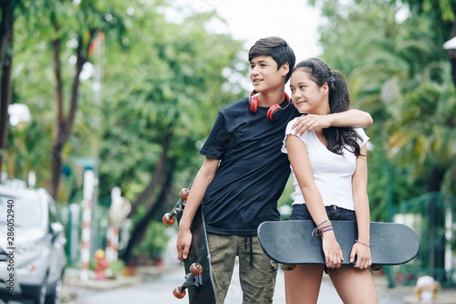 Cheerful hugging mixed-race brother and sister standing outdoors with skateboards in hands