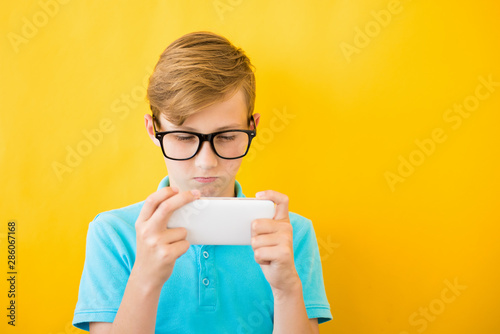 Handsome boy in glasses plays tablet on yellow background.. The concept of poor eyesight, harm of gadgets, myopia