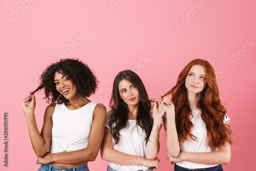 Thinking thoughtful young three multiethnic girls friends posing isolated over pink wall background.