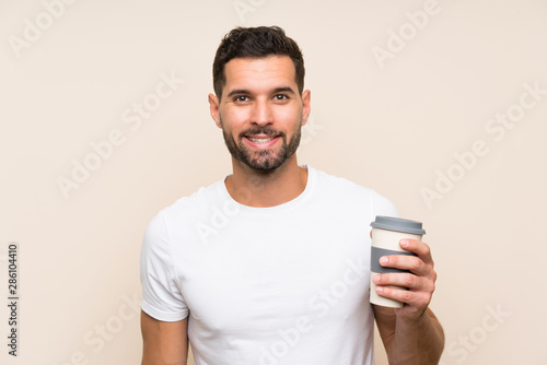 Young man with beard holding a take away coffee over isolated blue background smiling a lot