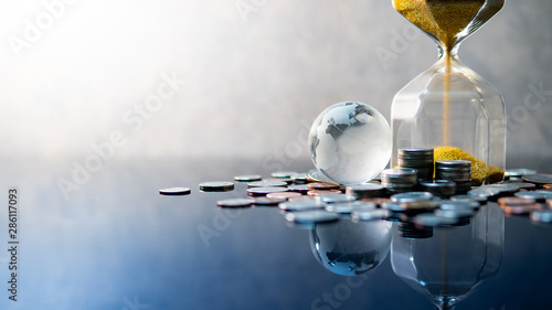 Global business investment growth. Wealth concept. Gold sand running through the shape of modern hourglass with world globe crystal glass and US dollar coins on blue reflective table.