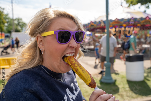 Blonde woman eats a pronto pup corn dog covered in mustard at an outdoor fair