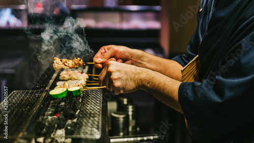 Hands of Japanese Yakitori Chef grilling chicken marinated with ginger, garlic and soy sauce and cucumber with a lot of smoke.