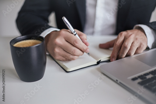 manos de hombre con traje escribiendo sobre libreta, con computadora y cafe