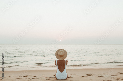 Summer vacation fashion concept. Young, tanned woman wearing a beautiful white swimsuit with a straw hat is sitting and relaxing on tropical beach with white sand and is watching sunset and sea.