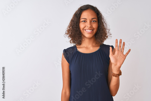 Young brazilian woman wearing blue dress standing over isolated white background showing and pointing up with fingers number five while smiling confident and happy.