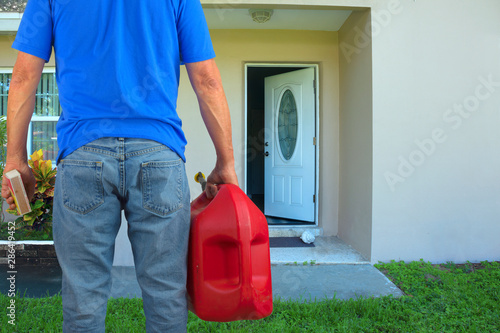 Arsonist man with red plastic gasoline can container and box of striking matches preparing to commit arson crime and maliciously and intentionally burn down a house with an open front door.