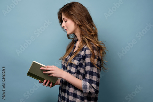 A young girl in a plaid shirt stands in profile on a blue background and looks down at the book with his head bowed.