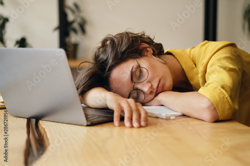 Young tired woman in eyeglasses sleeping on desk with laptop at workplace alone