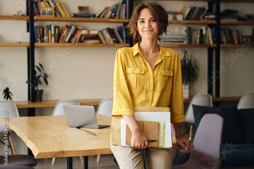 Young beautiful woman in yellow shirt leaning on desk with notepad and papers in hand while happily looking in camera in modern office